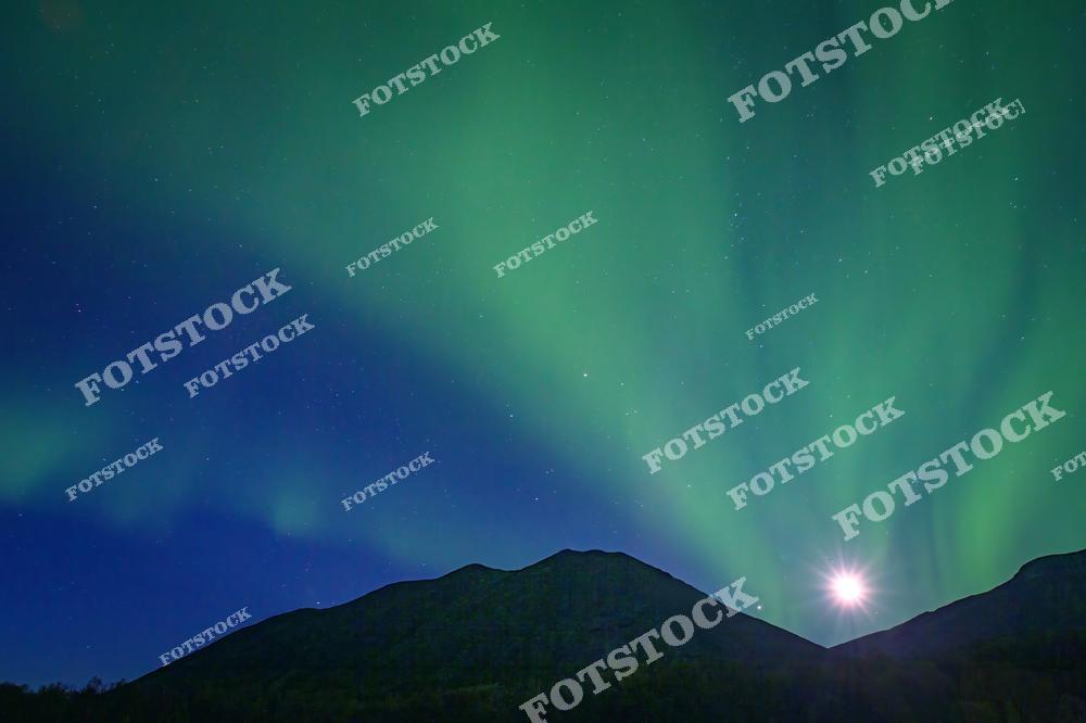 На фотографии изображено полярное сияние, освещающее небо над Хибинами. Внизу видны горы, а в центре кадра — свет от луны, который добавляет дополнительное сияние. Это типичное природное явление для Кольского полуострова и северных регионов России.